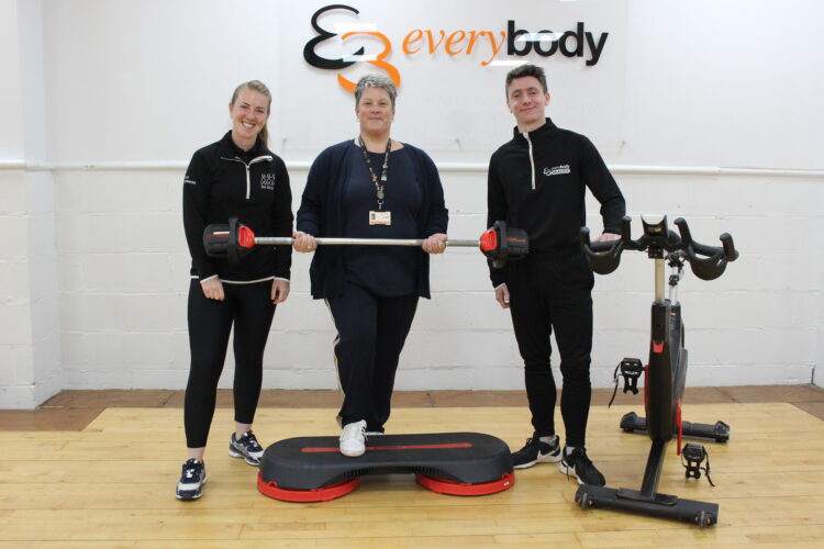 A group photo which includes, from left to right, Lucy Holmes, Wellbeing Lead at One Wirral CIC and the Manager of the Cancer Prehabilitation Service, Donna Williamson Senior Manager at Everybody Health and Leisure and Gavin McKeith Health Referral Lead at Everybody Health and Leisure. The team are in the class studio at Holmes Chapel Community Centre, with a variety of equipment such as, a group cycling bike, step box and barbell. 