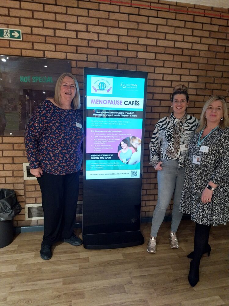 Image of Debbie Sharred Motherwell Wellbeing Coordinator with facilitators Natalie Coomer and Lisa Moss standing next to the digital screen at Macclesfield Leisure Centre promoting the Menopause Cafe