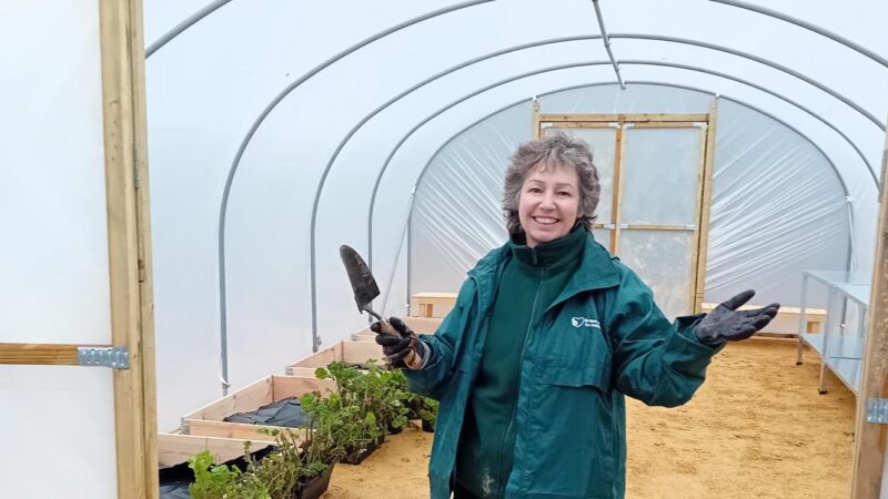 Green Spaces for Wellbeing member of staff stood in the Polytunnel, where the allotment drop in sessions will take place. Holding a gardening trowel with flower beds behind her.