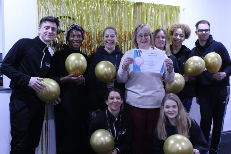Photo of the Everybody Healthy Team, standing in front of a gold backdrop, holding gold balloons and their certificate after being awarded ‘Recondition the Nation Gold Medal Award’ by NHS England.