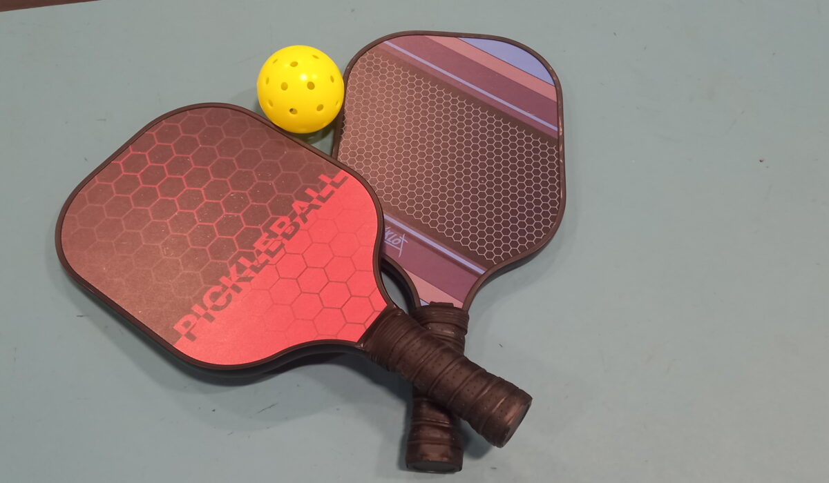 Image of two pickleball bats and a yellow ball on a green sports hall floor.