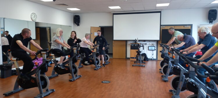 A group image of our customers taking part in our Bike Therapy for People Living with Parkinson’s dedicated group cycling class at Crewe Lifestyle Centre. The image includes class participants riding an indoor stationary bicycle and staff from Everybody Health and Leisure talking to customers.