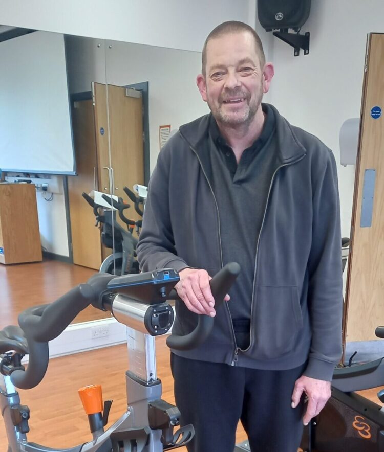 An image of Mike who attends our Bike Therapy for people living with Parkinson’s class. Mike is stood, smiling, next to one of our indoor stationary bicycles in our class studio at Crewe Lifestyle Centre