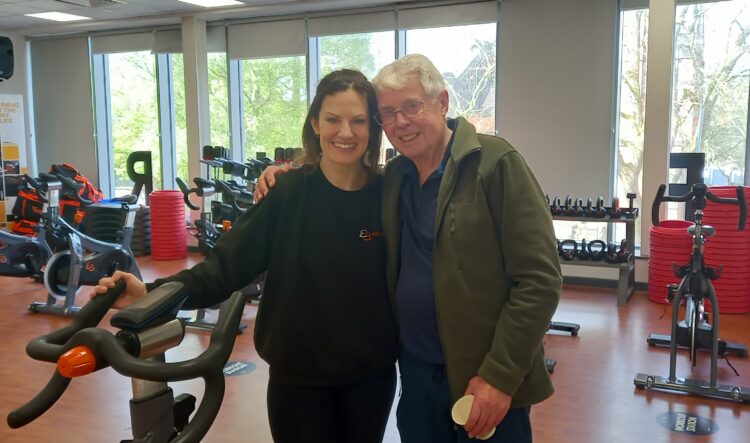 An image of Victoria Barker, Fitness Experience Manager and Bike Therapy for People Living with Parkinson’s class instructor at Everybody Health and Leisure, pictured on the Left. Victoria is stood next to Mike, another class participant. The photo is in the class studio at Crewe Lifestyle Centre, both Victoria and Mike are smiling and have indoor stationary bicycles in the background of the photo.