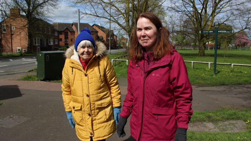 An image taken at the Ramblers Wellbeing Walk in Nantwich of two ladies stood smiling. They are stood on the pavement with houses and grass in the background.