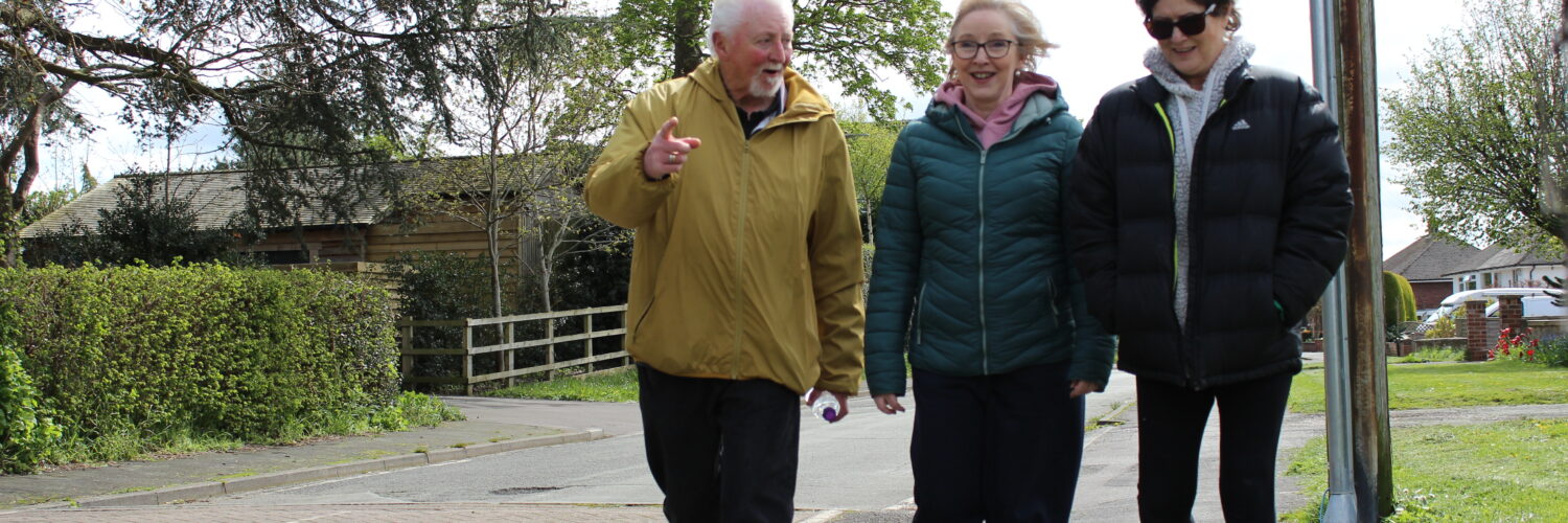A photo of three participants at the Ramblers Wellbeing Walk in Nantwich. The group are walking on the pavement down a street, smiling, pointing and chatting.