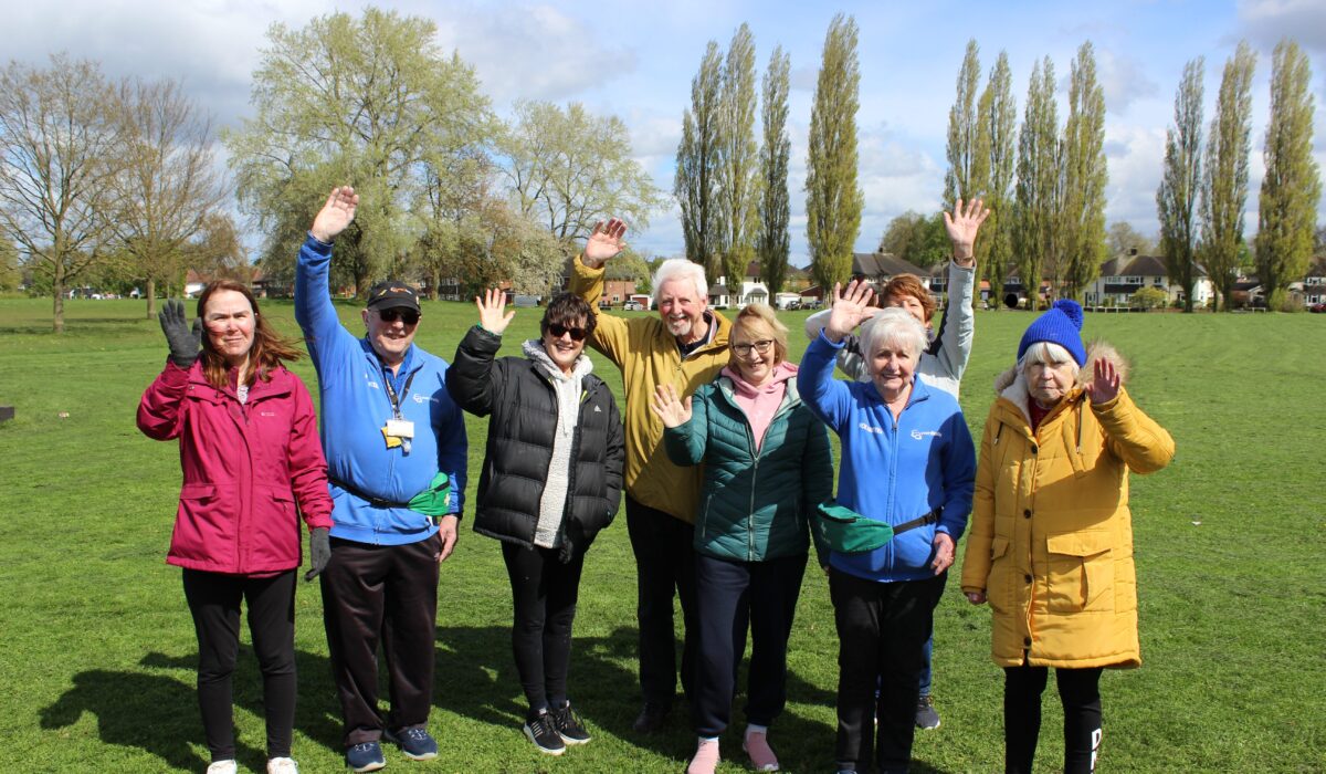 A group photo of participants and Everybody Volunteers at the Ramblers Wellbeing Walk in Nantwich. The group are stood in a field, with trees in the background, on a sunny day with clouds in the sky. Participants are smiling and waving at the camera.