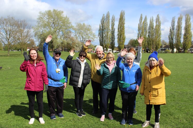 A group photo of participants and Everybody Volunteers at the Ramblers Wellbeing Walk in Nantwich. The group are stood in a field, with trees in the background, on a sunny day with clouds in the sky. Participants are smiling and waving at the camera.