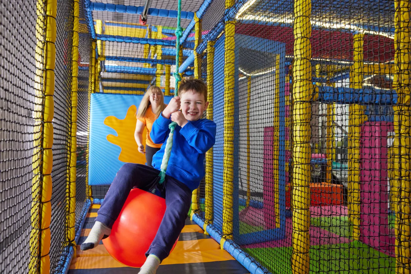 A mother pushes her son on a fishing bouy zipline in a brightly coloured kids softplay