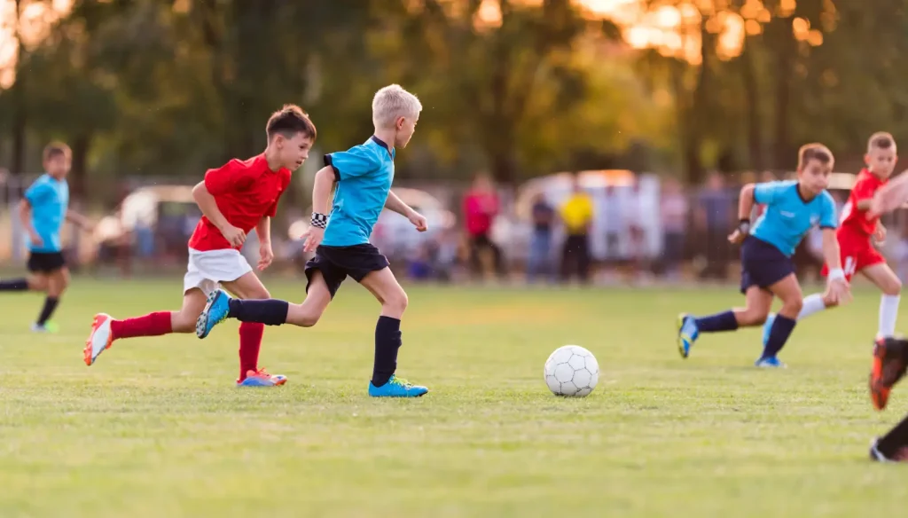 Young children in red and blue football kits chasing the ball during a match on a grass pitch at sunset