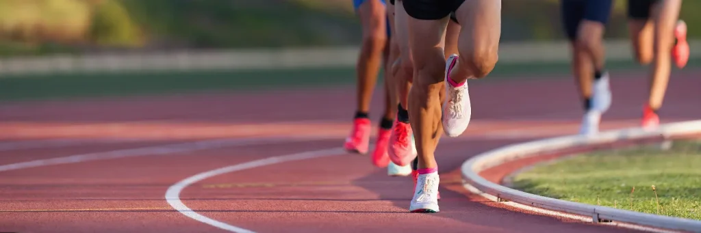 Runners competing on a red athletics track, photographed at ground level showing their legs and running shoes as they round a bend